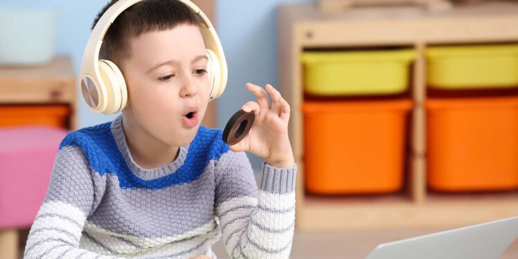 Ein Kind, das Kopfhörer trägt und vor einem Laptop sitzt, hat den hözernen Buchstaben O in der linken Hand und formt mit dem Mund den Laut O.