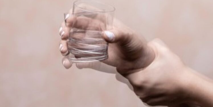 Eine menschliche Hand hält ein Glas Wasser. Das Bild ist leicht verschwommen und deutet eine zittrige Handbewegung an.